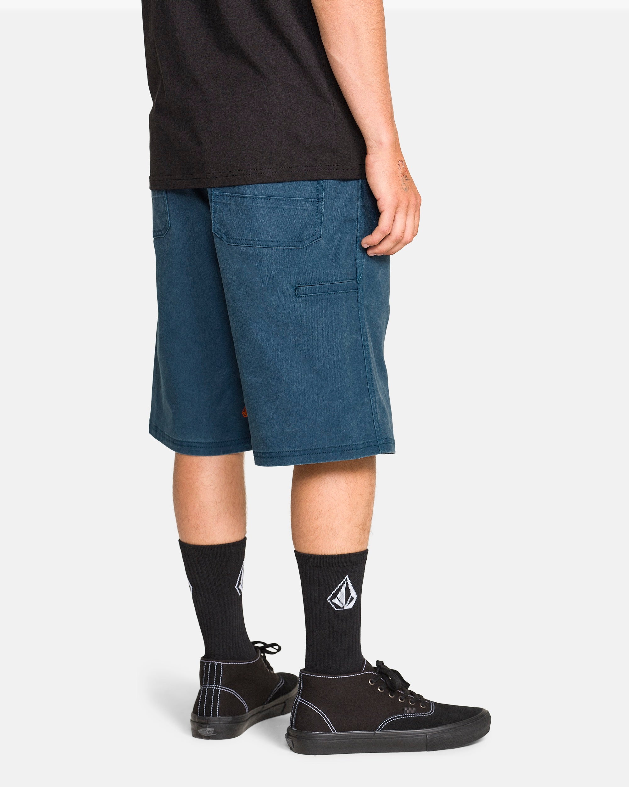 A man in a black T-shirt, Volcom Men's Dialed Relaxed Work Shorts in teal, black socks with a white logo, and black sneakers stands against a plain white background, viewed from the side and slightly from the back.