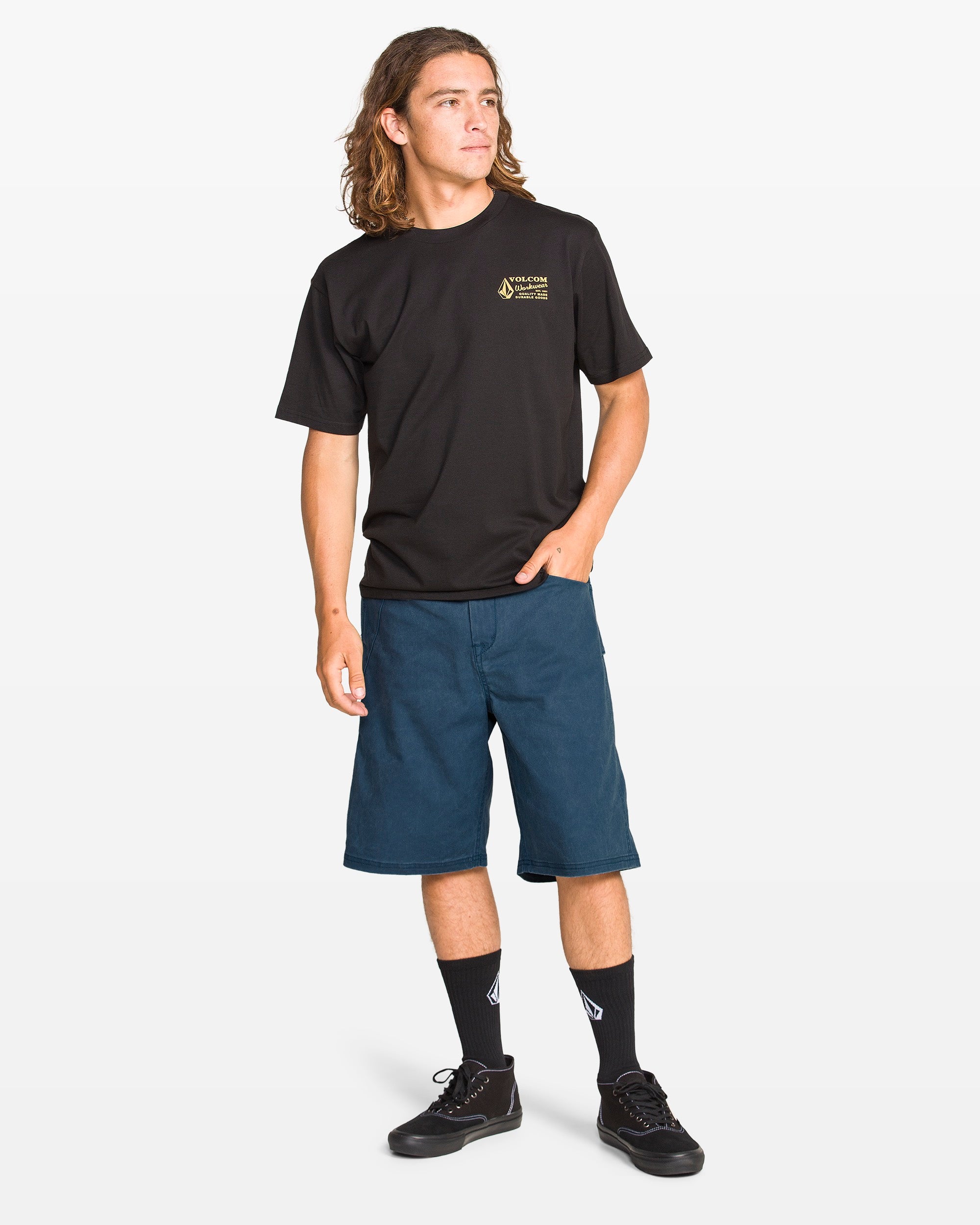 A young man with long hair wears a black T-shirt, Volcom Men's Dialed Relaxed Work Shorts in blue, black socks, and sneakers. He stands with one hand in his pocket, looking to the side against a plain white background.