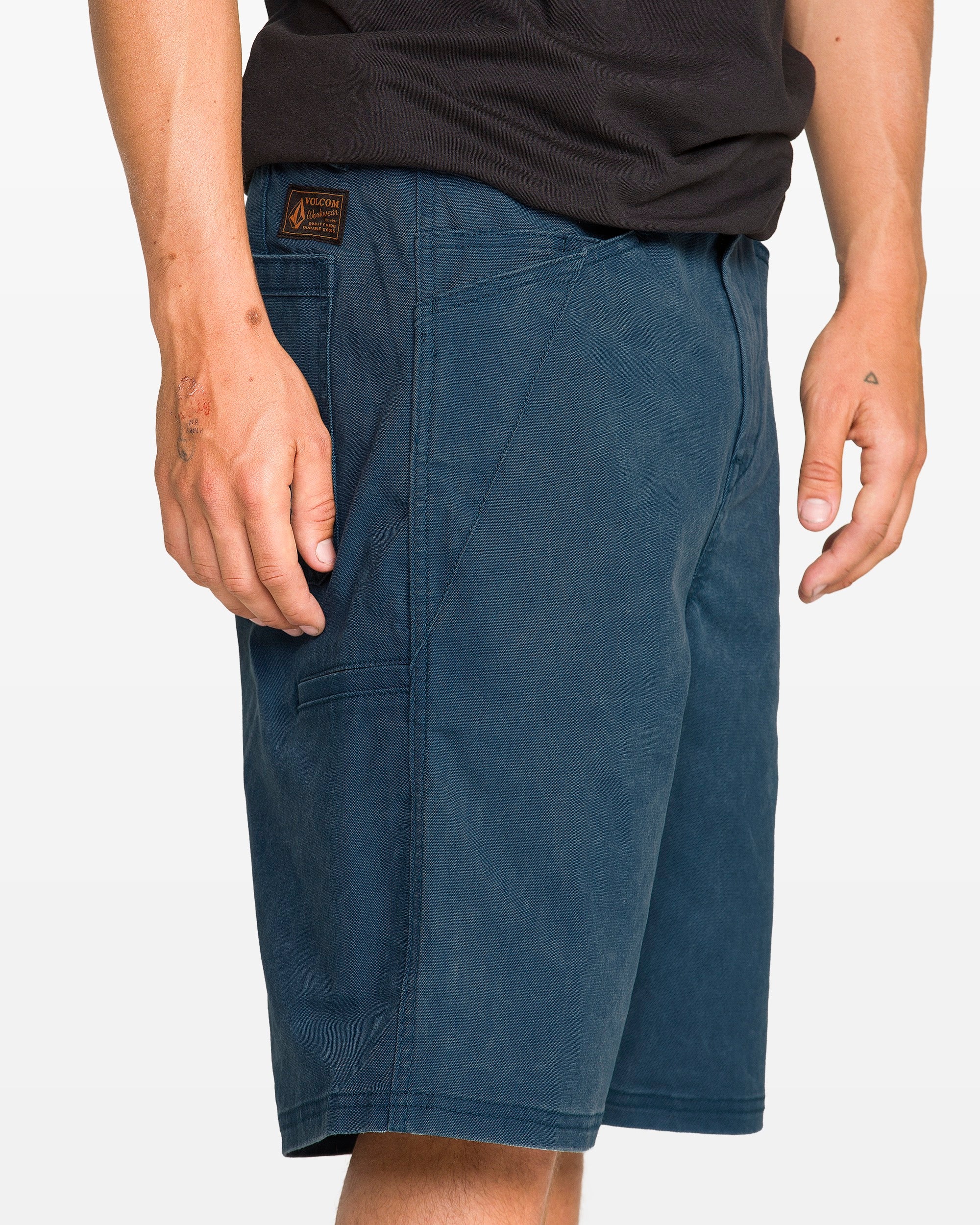 A person is shown from waist to knee, wearing Volcom Men's Dialed Relaxed Work Shorts in blue with a visible patch near the pocket, paired with a black T-shirt.