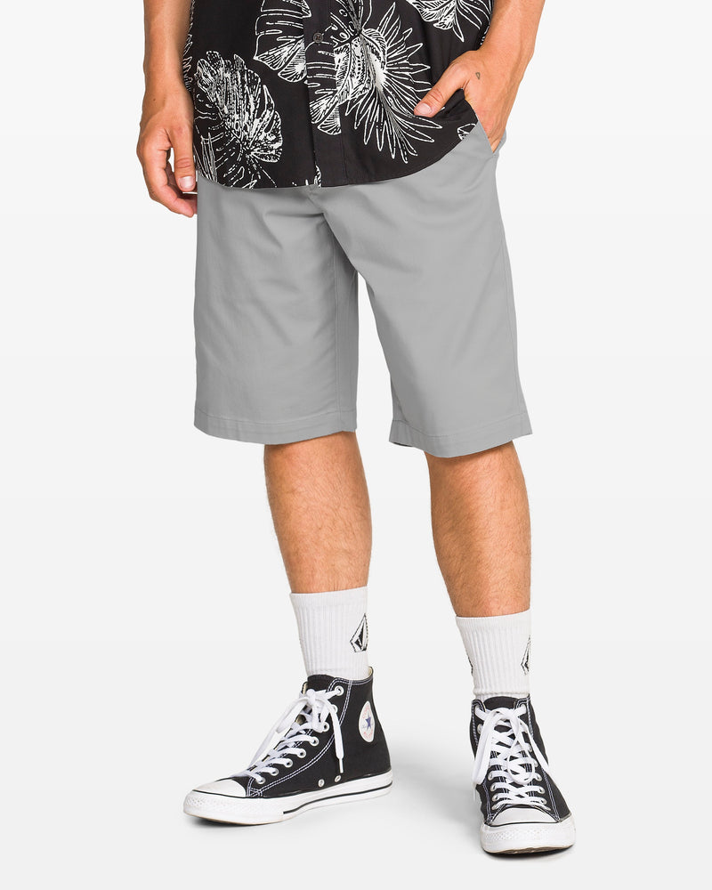 A person wearing a black and white patterned shirt, Volcom Men's Frickin Modern Stretch Shorts, white socks, and black high-top Converse sneakers stands with one hand in their pocket.