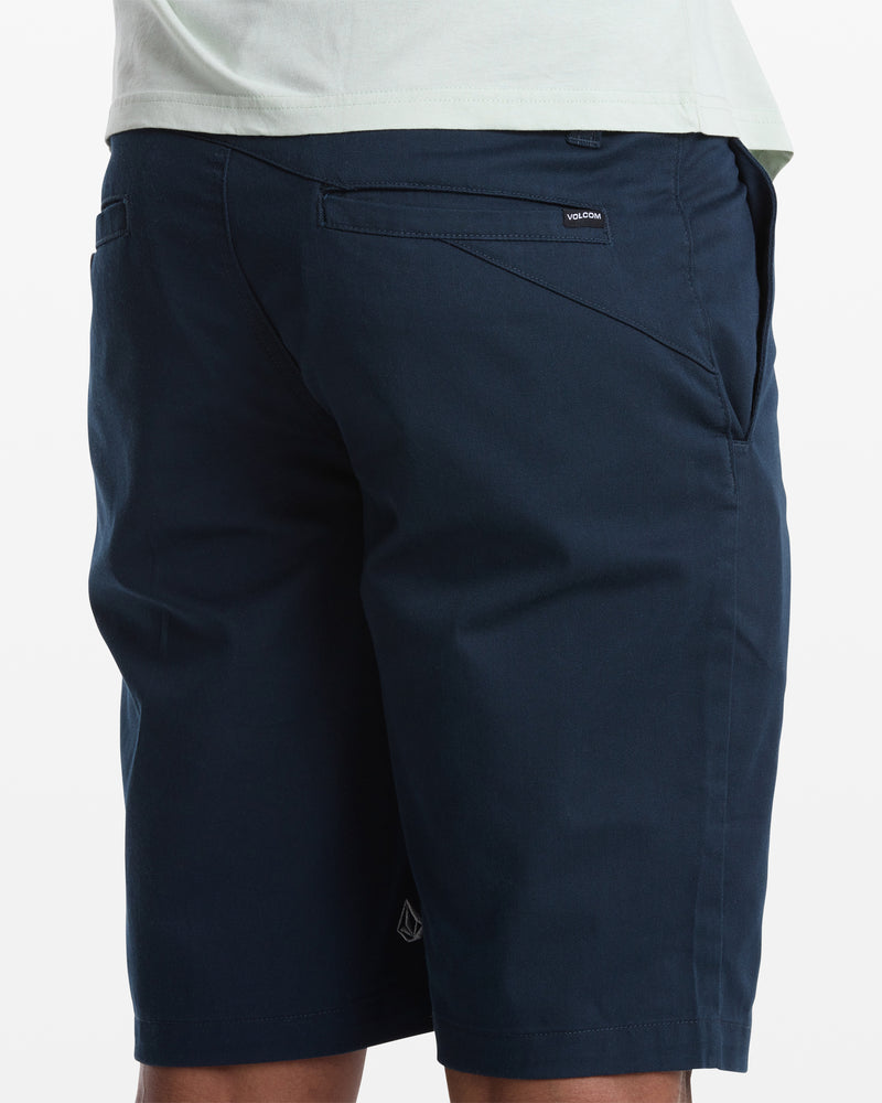 A person shown from the back, waist down, wears dark blue Volcom Men's Frickin Modern Stretch Shorts with pockets and a small black Volcom label above the right pocket, paired with a light-colored shirt.