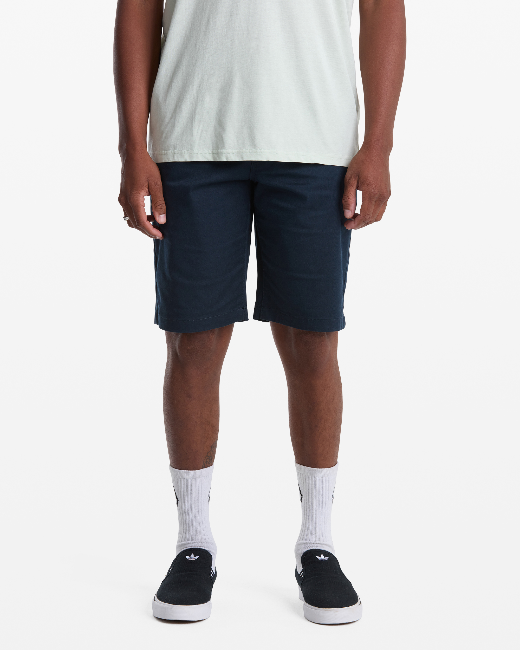 A person wearing a light T-shirt, Volcom Men's Frickin Modern Stretch Shorts in navy, white socks with black Adidas logos, and black slip-on shoes stands against a white background; the image is cropped at the shoulders.
