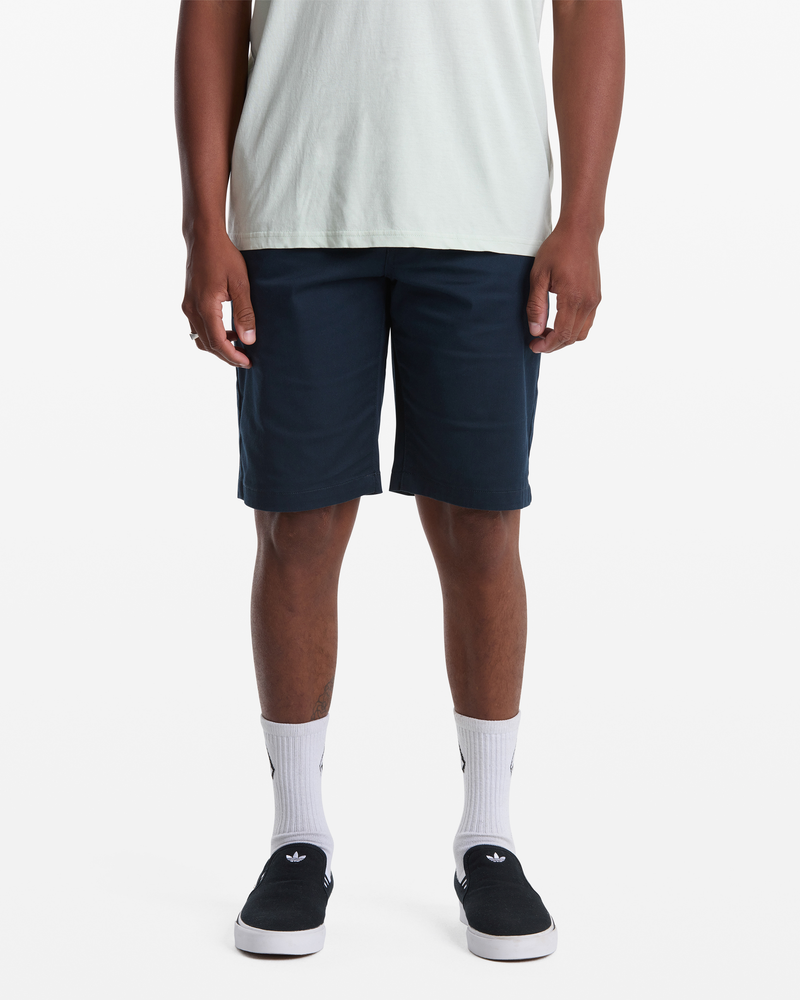 A person wearing a light T-shirt, Volcom Men's Frickin Modern Stretch Shorts in navy, white socks with black Adidas logos, and black slip-on shoes stands against a white background; the image is cropped at the shoulders.