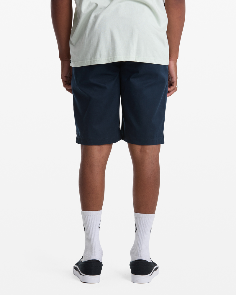 A person stands facing away against a white background, wearing a light T-shirt, Volcom Men's Frickin Modern Stretch Shorts in navy blue, white crew socks, and black slip-on shoes.