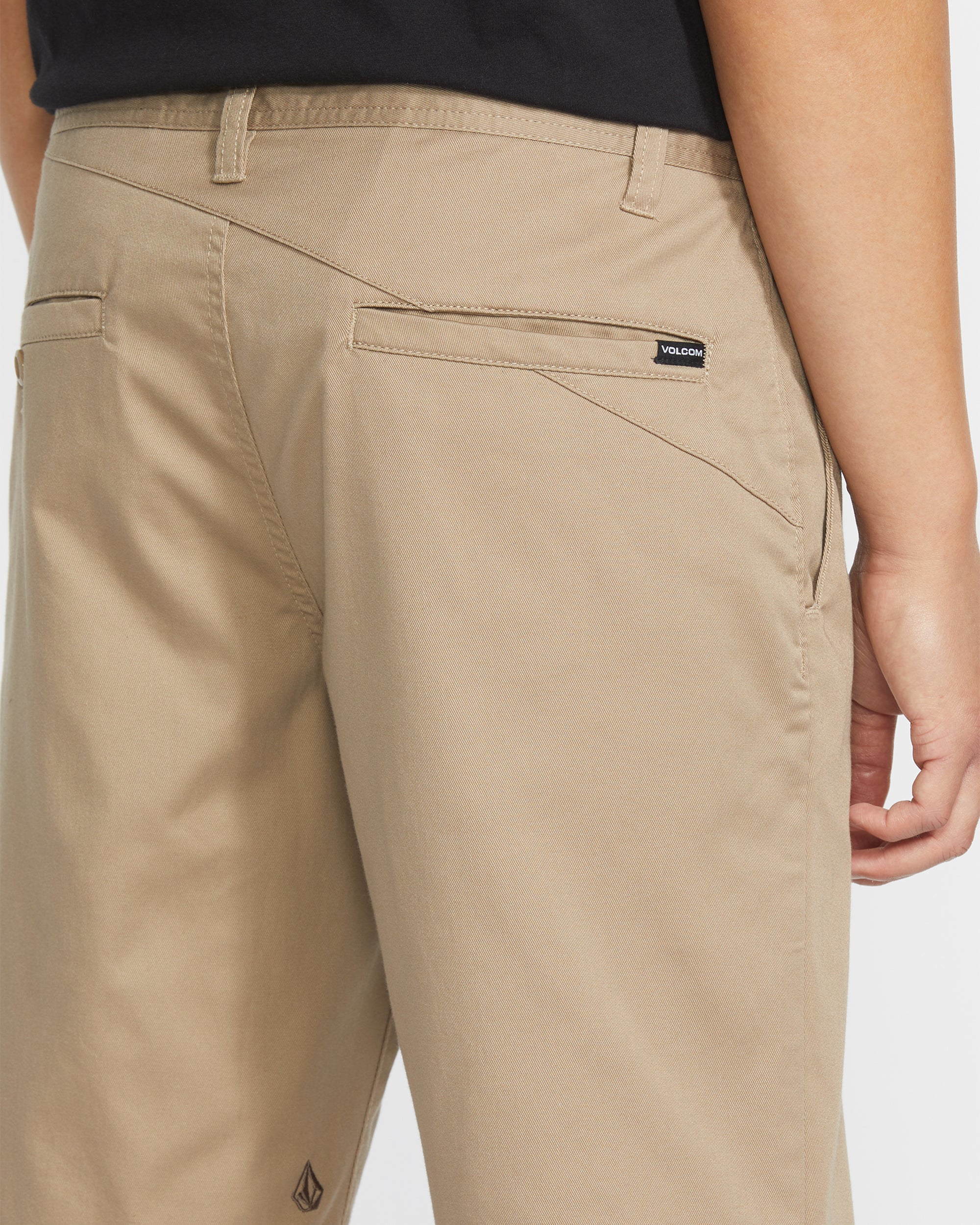 A close-up of the back of a person wearing tan Volcom Men's Frickin Modern Stretch 21" Shorts and a black shirt, highlighting the pocket details and the Volcom brand tag above the right back pocket.