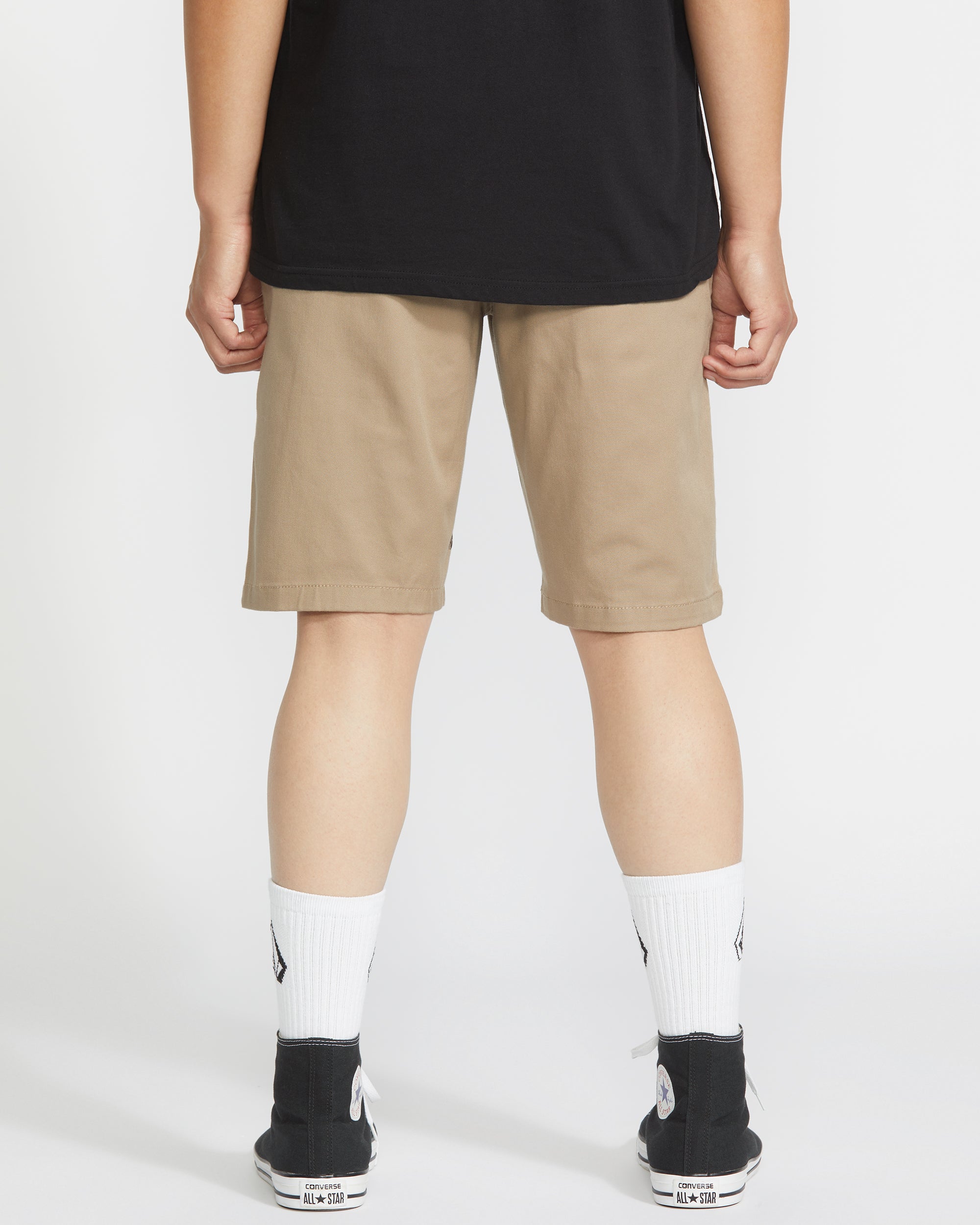 A person stands facing away from the camera wearing a black shirt, beige Volcom Men's Frickin Modern Stretch 21" Shorts, white socks, and black sneakers against a white background.