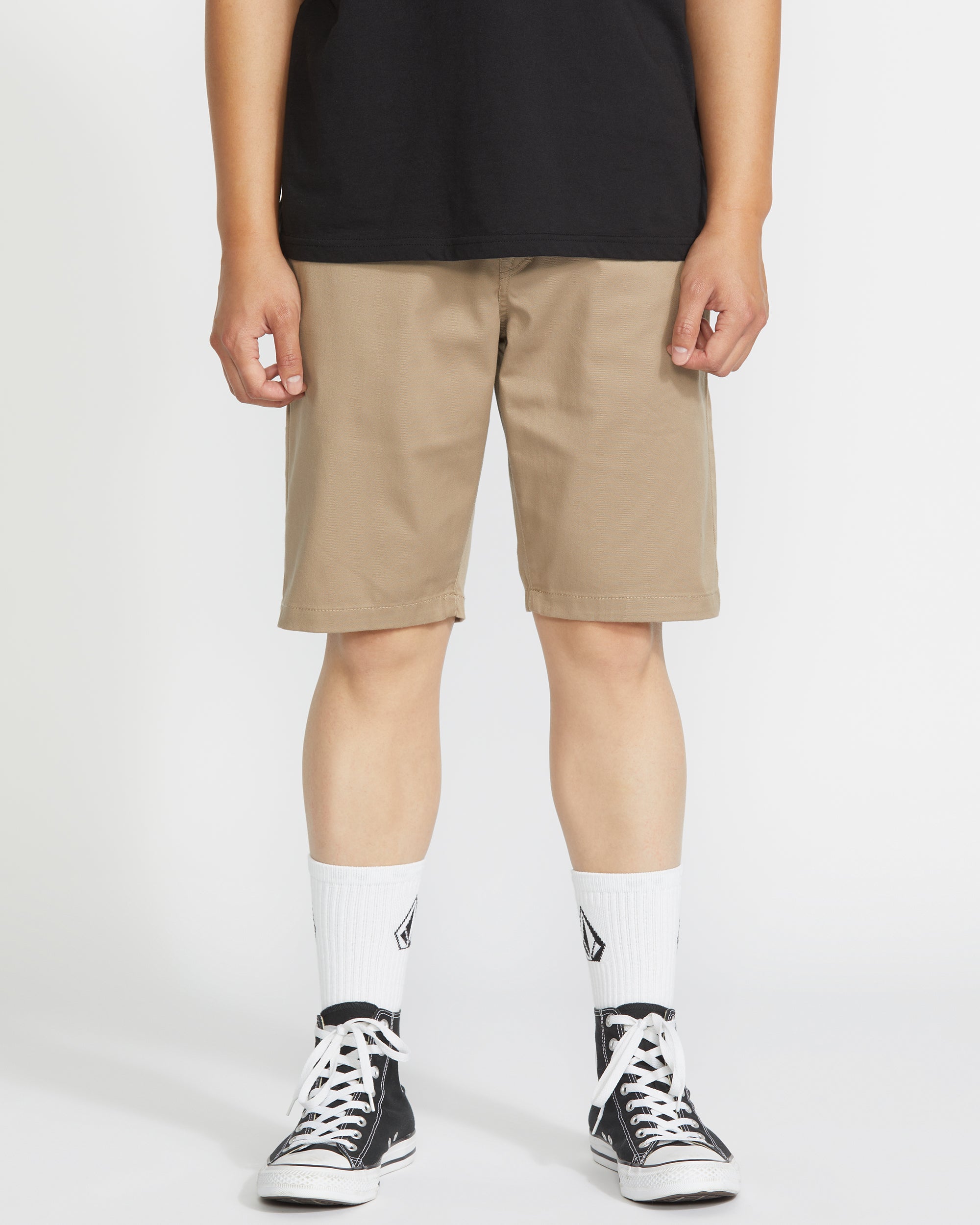 A person stands against a plain white background, wearing a black t-shirt, Volcom Men's Frickin Modern Stretch 21" shorts in khaki, white crew socks with a black logo, and black low-top sneakers.