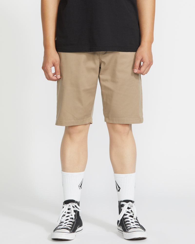 A person stands against a plain white background, wearing a black t-shirt, Volcom Men's Frickin Modern Stretch 21" shorts in khaki, white crew socks with a black logo, and black low-top sneakers.