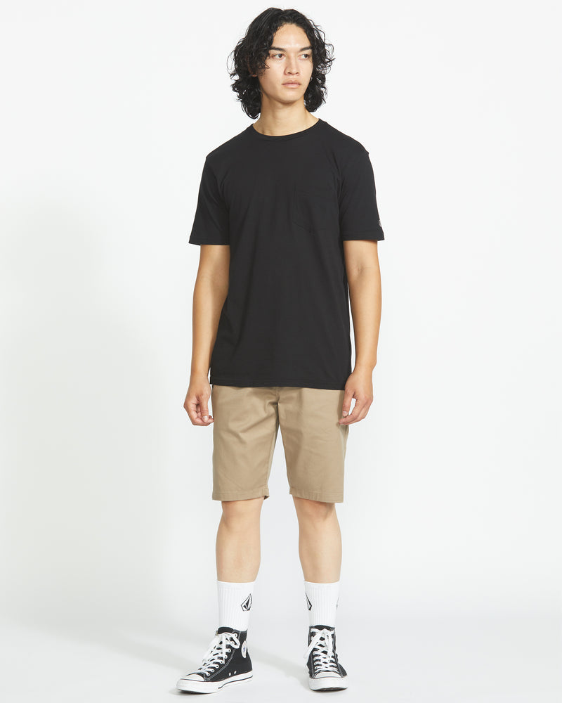 A person with curly hair wears a black t-shirt, Volcom Men’s Frickin Modern Stretch 21" Shorts in khaki, white socks with a black logo, and black Converse sneakers, standing against a plain white background.