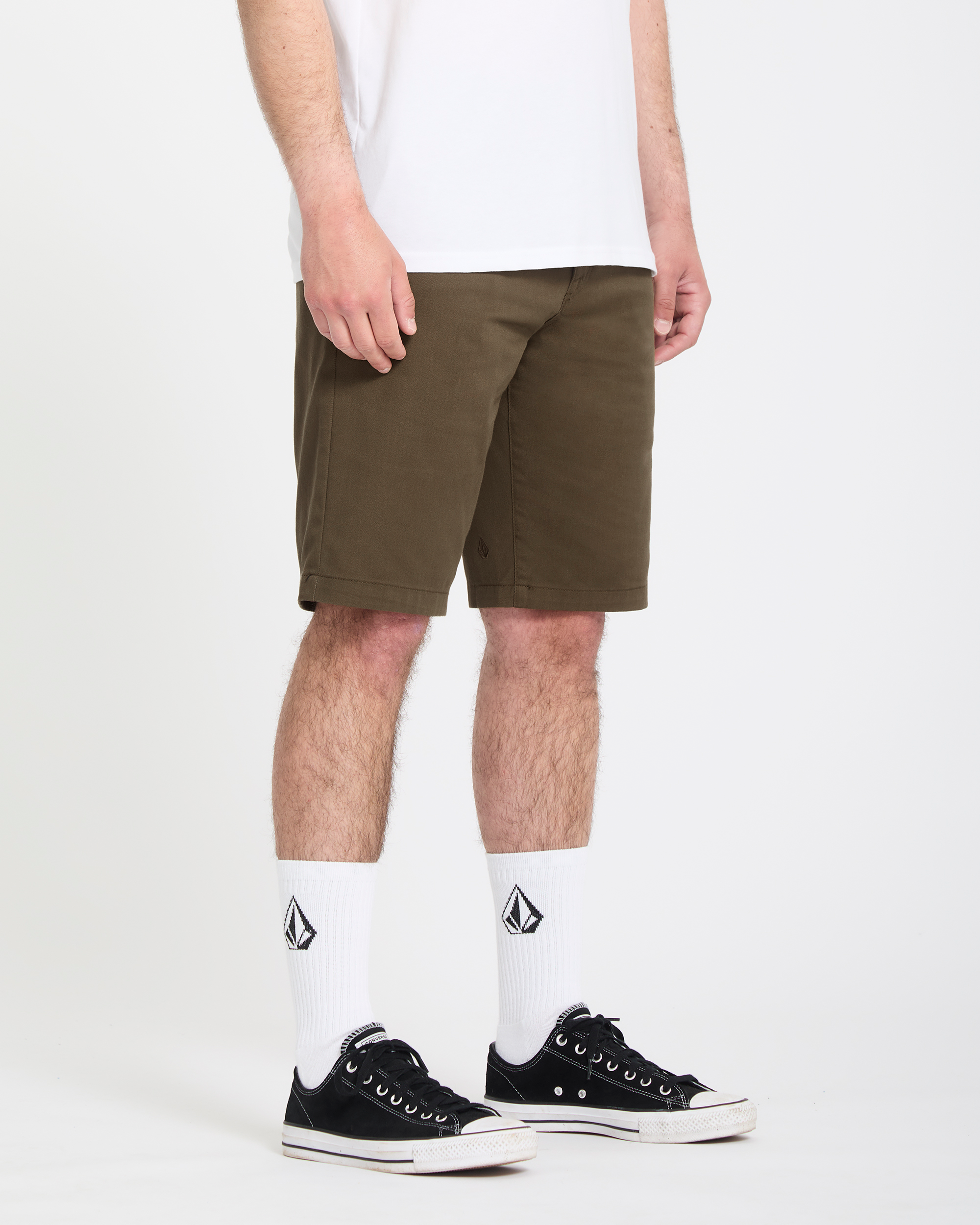 A person wearing a white t-shirt, Volcom Men's Frickin Modern Stretch Shorts in olive green, white crew socks with a black logo, and black low-top sneakers stands against a plain white background.