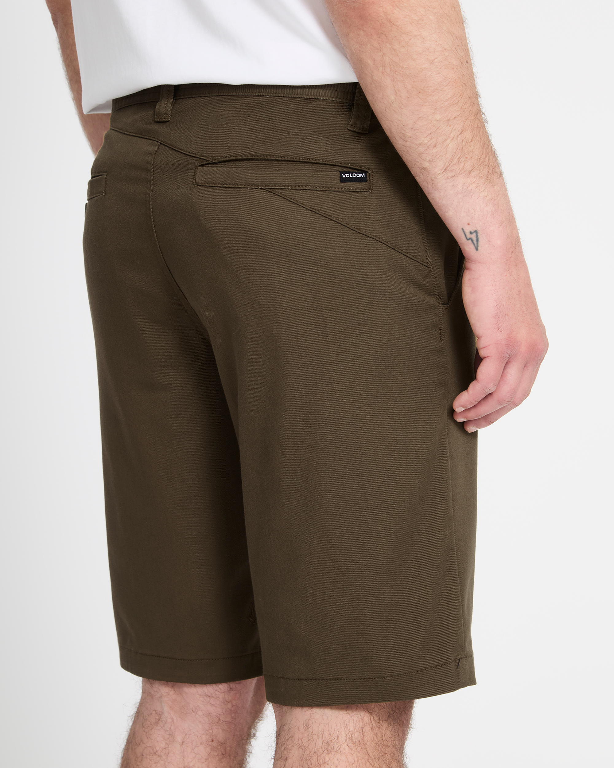 A person shown from the back wears Volcom Men's Frickin Modern Stretch Shorts in olive green with back pockets and a black label on the waistband, paired with a white shirt. Their right arm is visible, displaying a small tattoo.