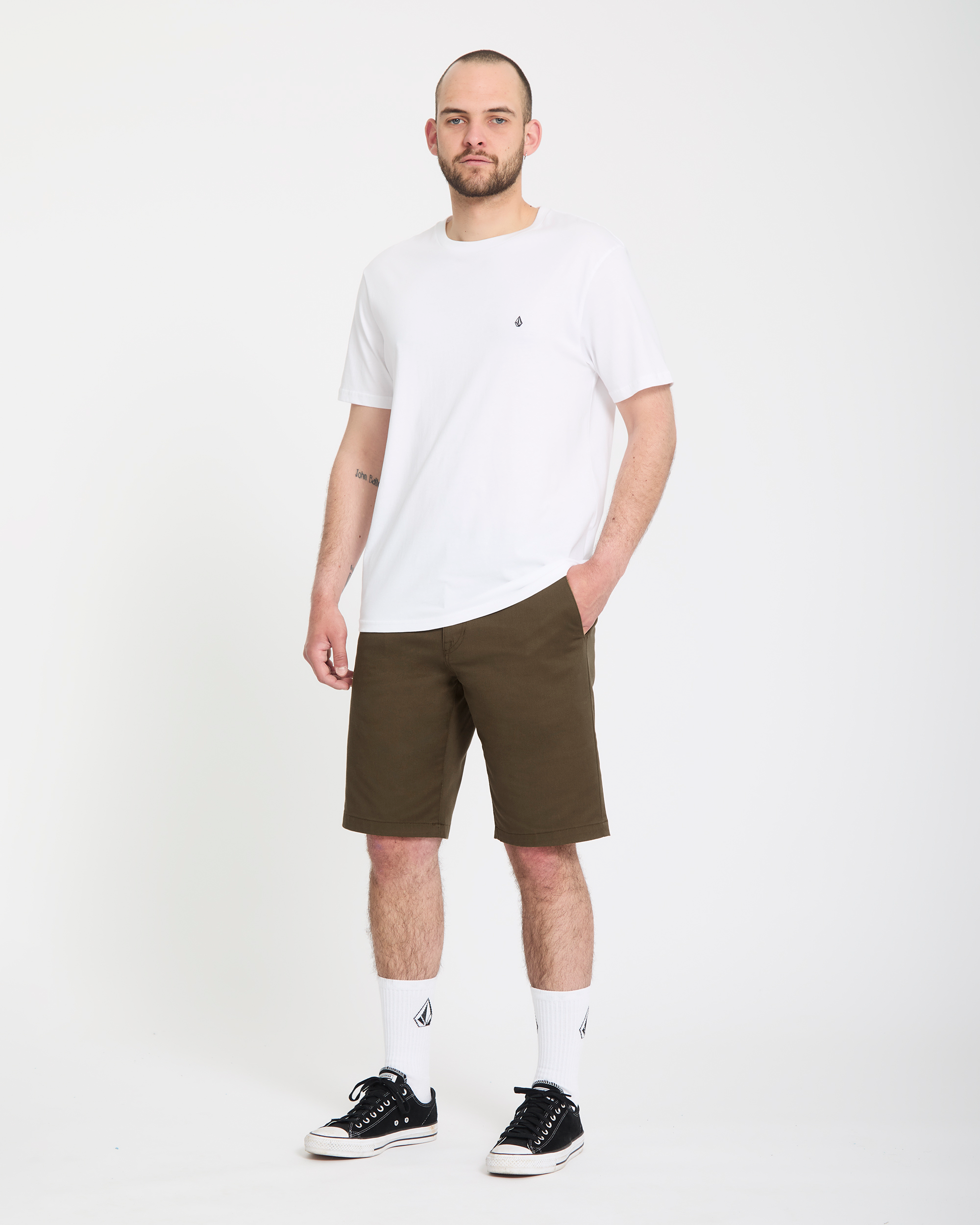 A man with a shaved head and beard wears a white tee, white socks, black sneakers, and Volcom Men's Frickin Modern Stretch Shorts in olive green, standing against a plain white background with a neutral expression.