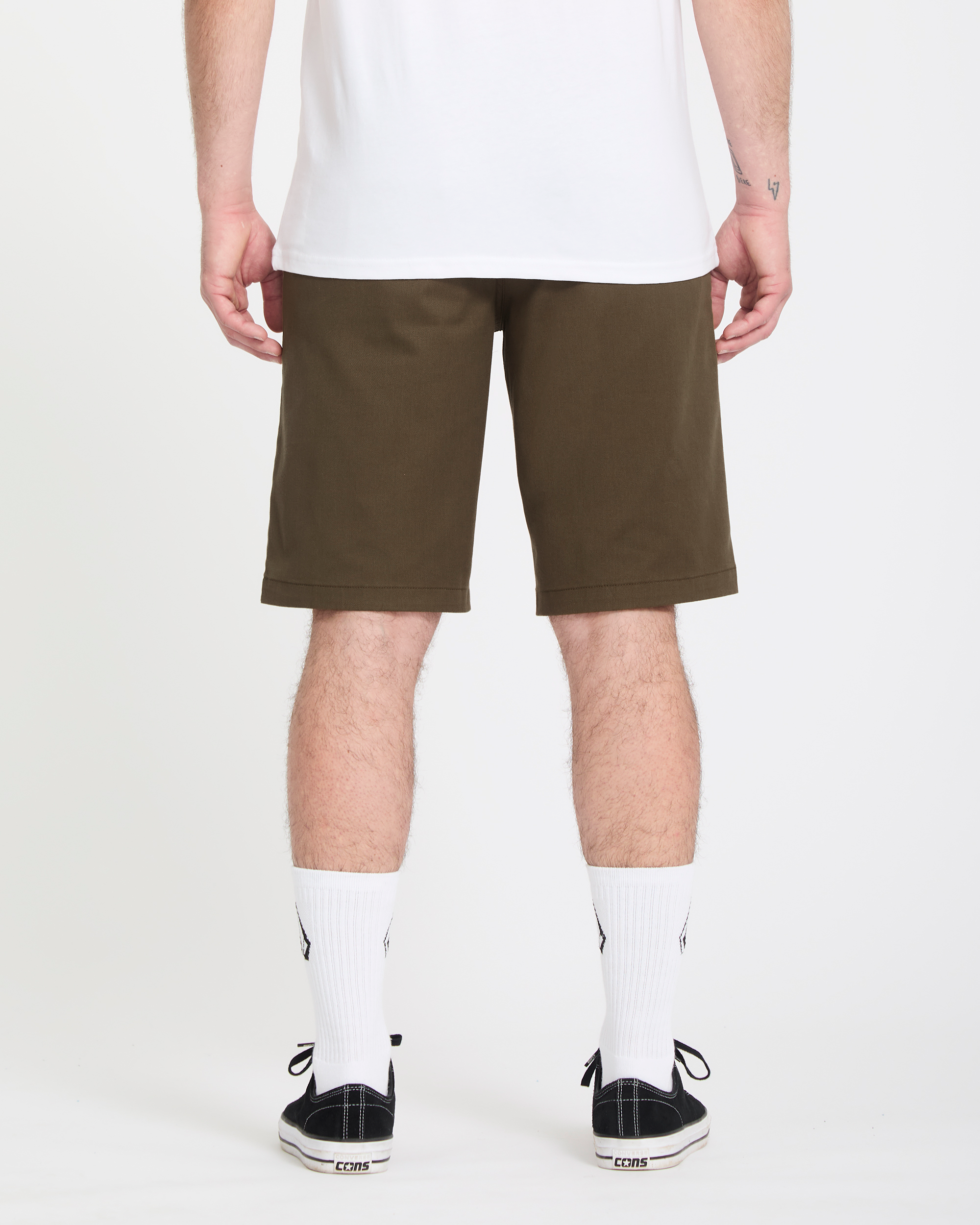 A person faces away from the camera wearing a white tee, Volcom Men's Frickin Modern Stretch olive green shorts, white crew socks, and black sneakers with white soles, against a plain white background.