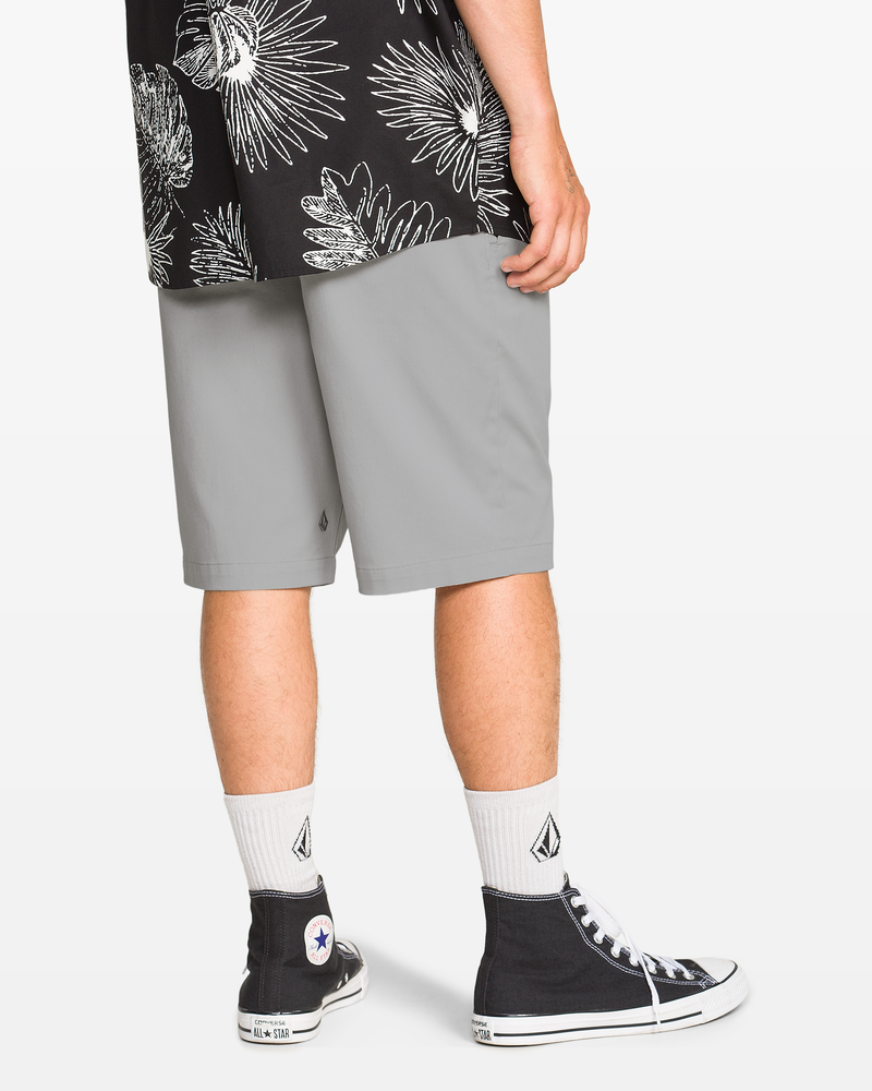A person stands facing away, wearing a black shirt with white floral print, Volcom Men's Frickin Modern Stretch Shorts in light gray, white socks with a black logo, and black high-top Converse sneakers.