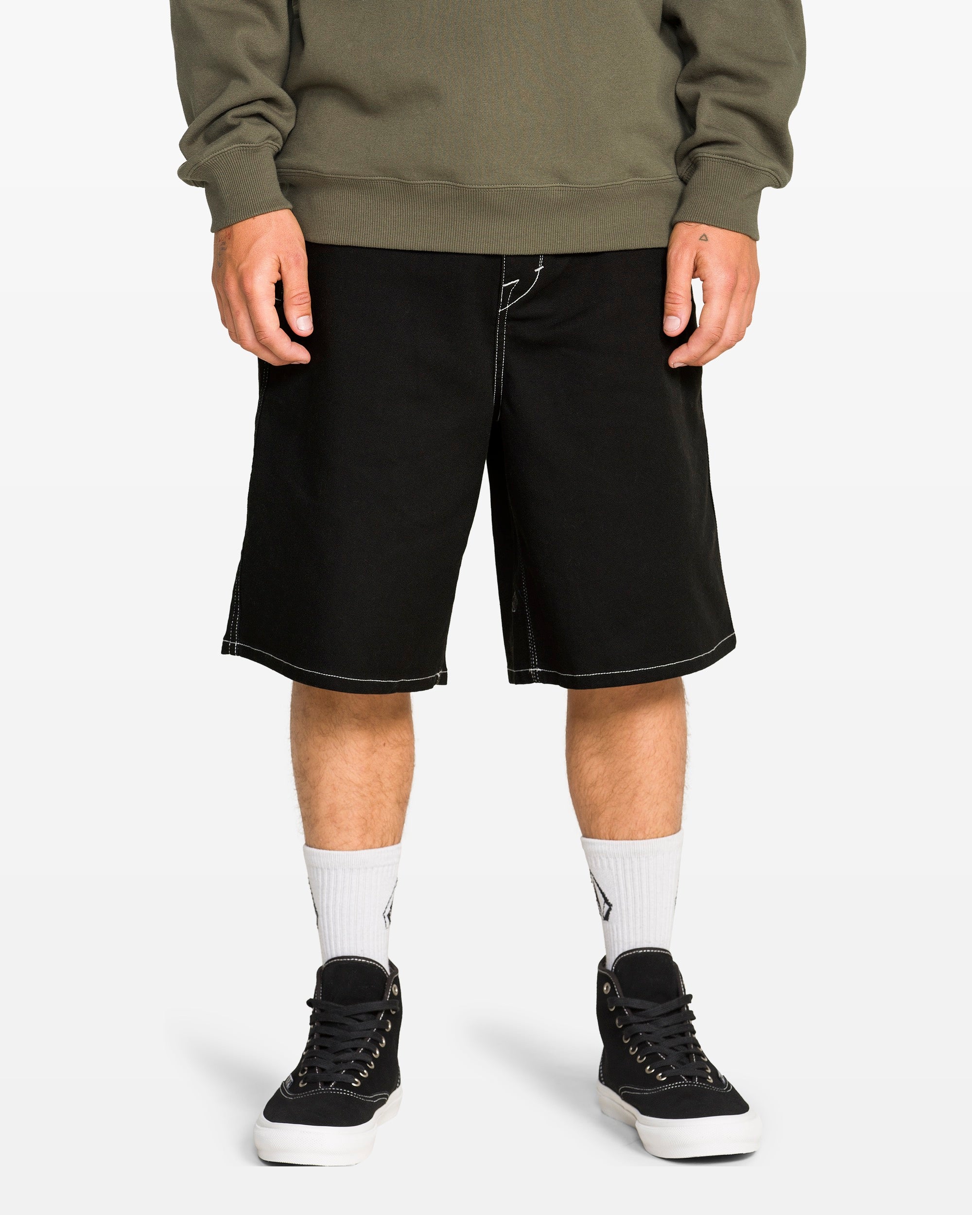 A person wears Volcom's Men's Billow Shorts in black twill with white stitching, white socks, black sneakers, and an olive green sweatshirt. Only the lower half is visible against a plain background. Shorts have a 22-inch outseam.