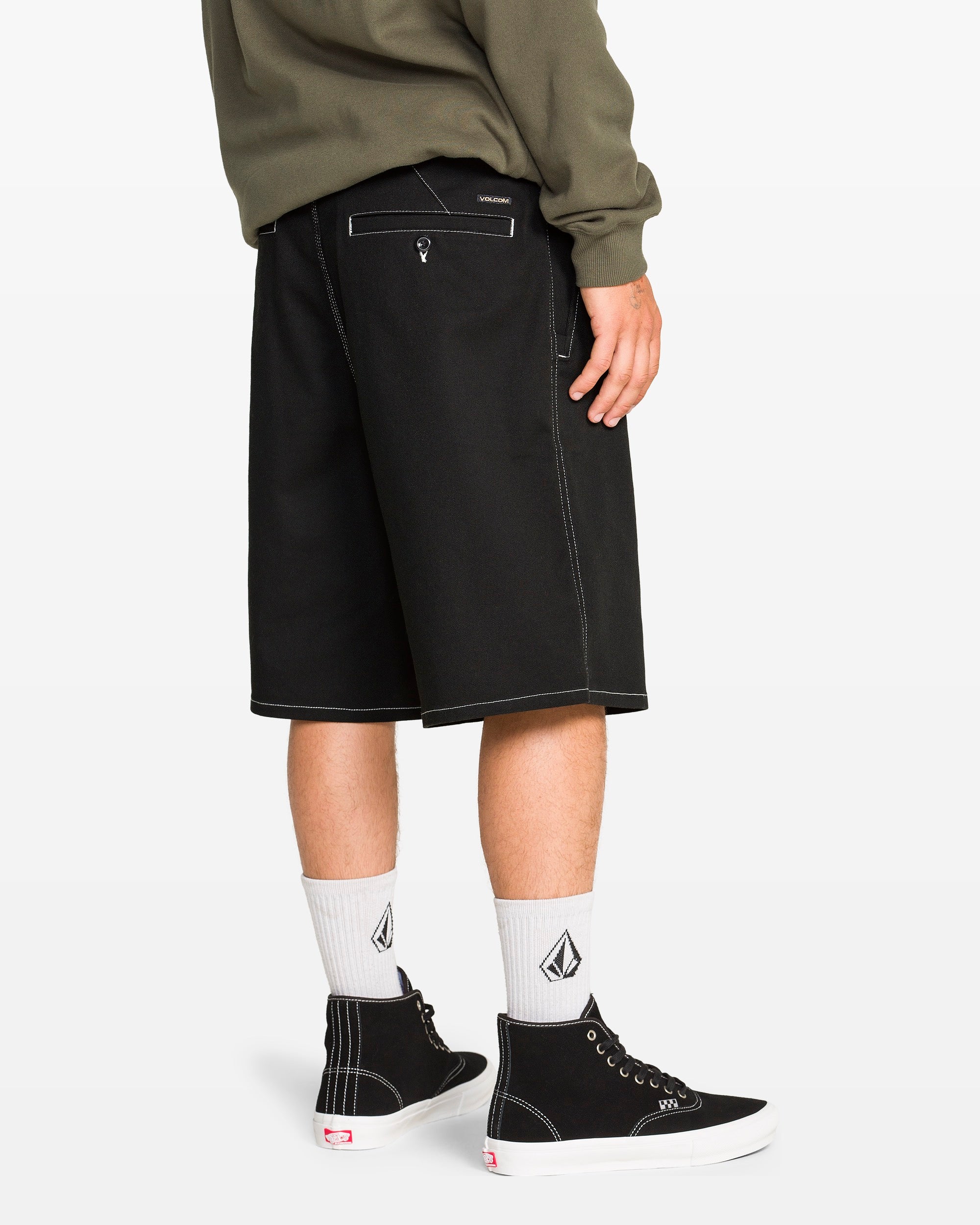 A person in Volcom Men's Billow Shorts in black (22" outseam), white socks with a black geometric logo, black high-top sneakers, and an olive green sweatshirt stands against a plain white background.