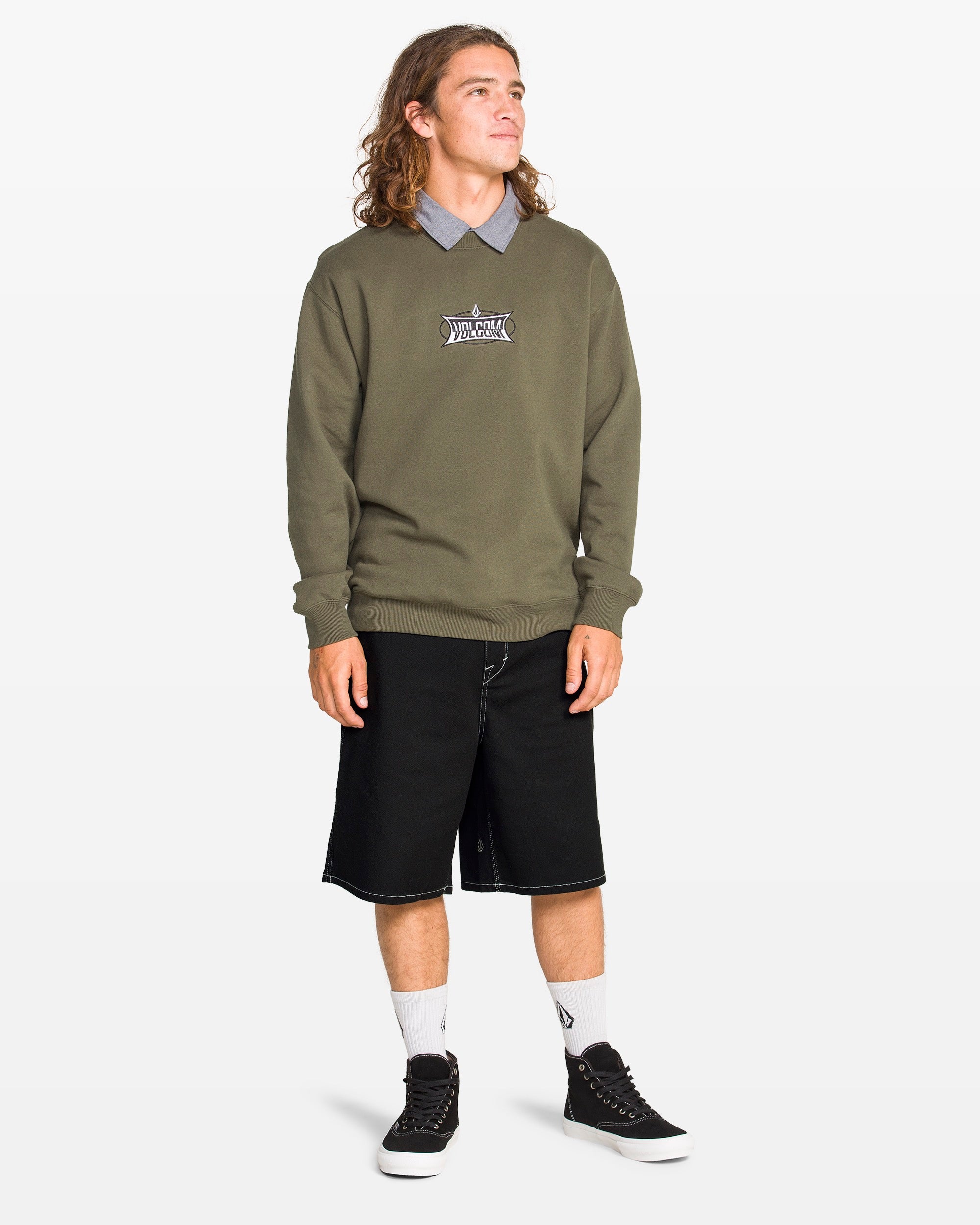 A person with long hair stands slightly to the side, wearing a green sweatshirt over a collared shirt, Volcom Men's Billow Shorts in black twill (22-inch outseam), white socks, and black sneakers against a plain white background.