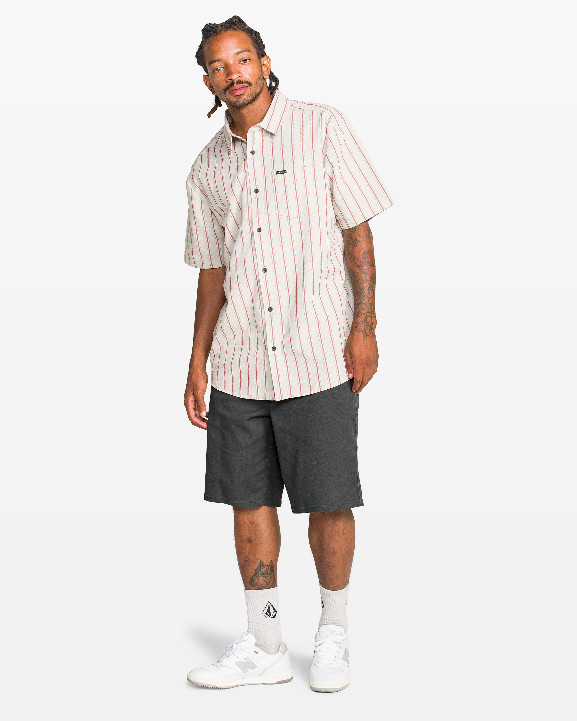 A man with braided hair and tattoos wears Volcom Men's Billow Short 22, a short-sleeve button-up white shirt with red stripes, white logo socks, and white sneakers, standing against a plain white background.