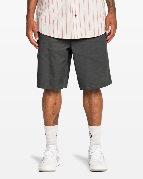 A person is shown from mid-chest down, wearing a white shirt with red stripes, white crew socks, white sneakers, and Volcom Men's Billow Short 22 in dark gray.