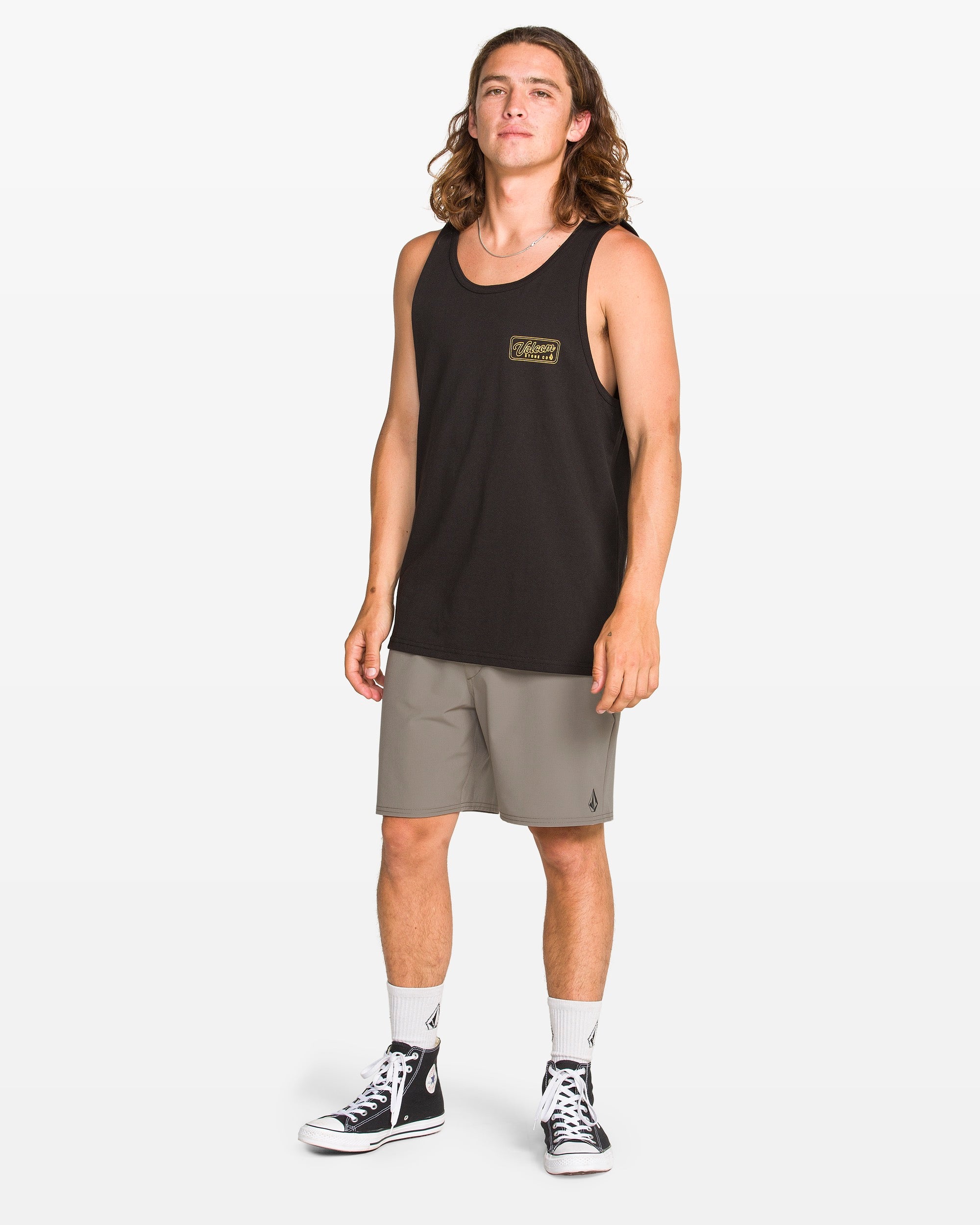 A person with long hair is wearing a black tank top, Volcom Men's Nomoly Elastic Waist Shorts 18", white crew socks, and black high-top sneakers, standing against a plain white background.