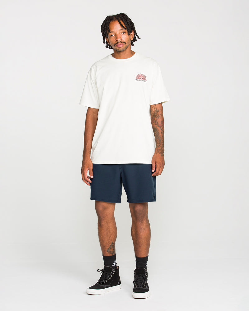 A man stands against a plain white background, wearing a white t-shirt with a small graphic on the chest and Volcom Men's Nomoly Elastic Waist Shorts in navy, which are made from a recycled blend. He completes his look with black socks and black high-top sneakers. He has tattoos on his arms and short, twisted hair.