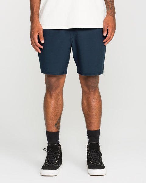 A person wearing a white shirt and Volcom Men's Nomoly Elastic Waist Shorts in dark blue, paired with black socks and black sneakers, stands against a plain background. Only the lower torso and legs are visible.