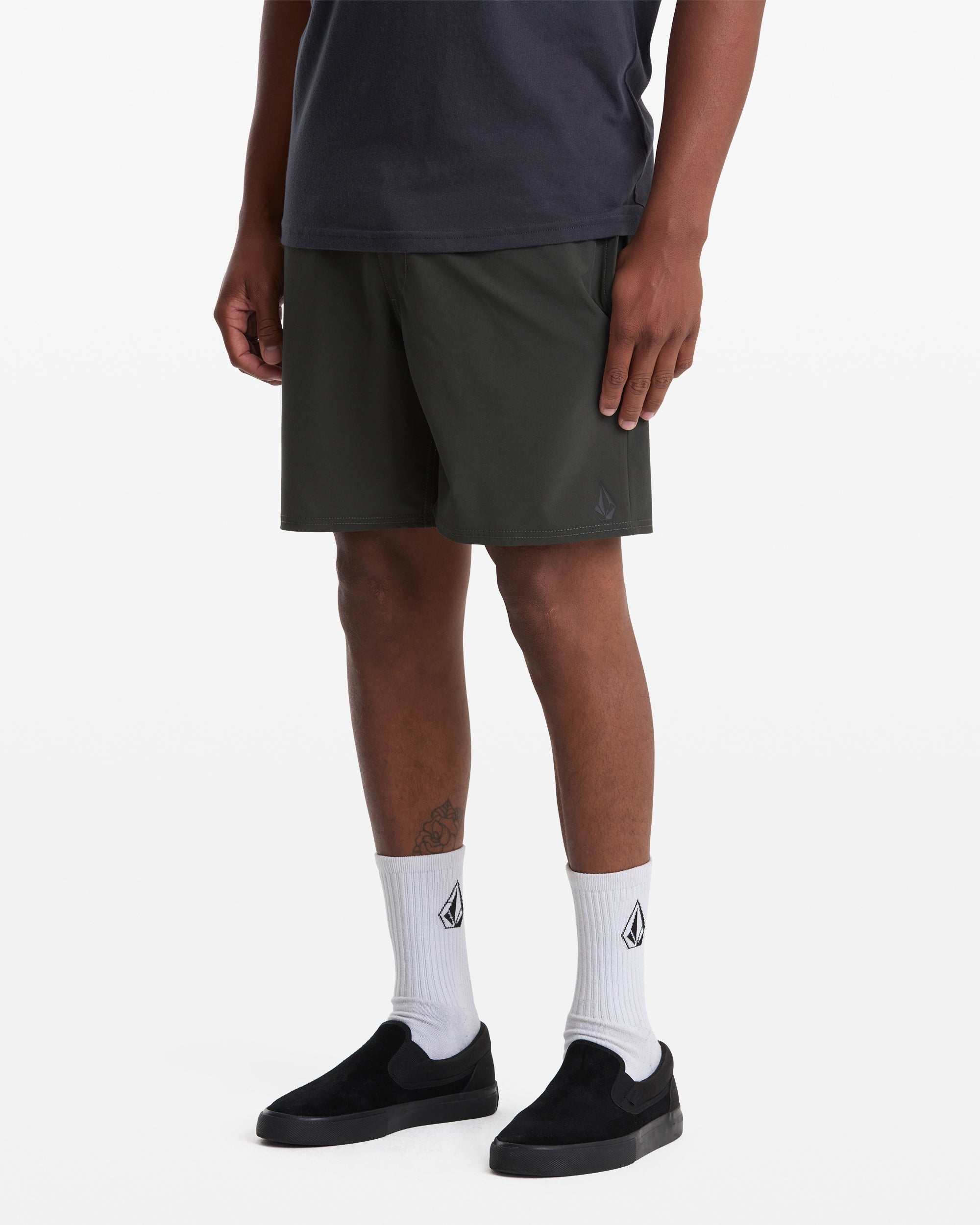 A person wearing a black t-shirt, Volcom Men's Nomoly Elastic Waist Short 18 in dark green, white socks with a logo, and black slip-on shoes, standing against a plain white background.