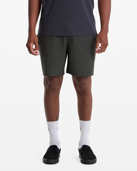 A person wears Volcom Men's Nomoly Elastic Waist Short 18 in dark gray, paired with a black t-shirt, white crew socks, and black slip-on shoes, standing against a plain white background. Image is cropped from the shoulders down.