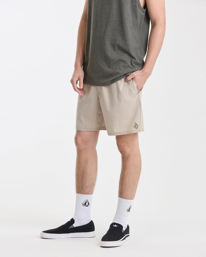 A man wearing a dark gray sleeveless shirt, Volcom Men's Nomoly Elastic Waist Short 18 in beige, white logo socks, and black slip-on shoes stands with one hand in his pocket against a plain white background.