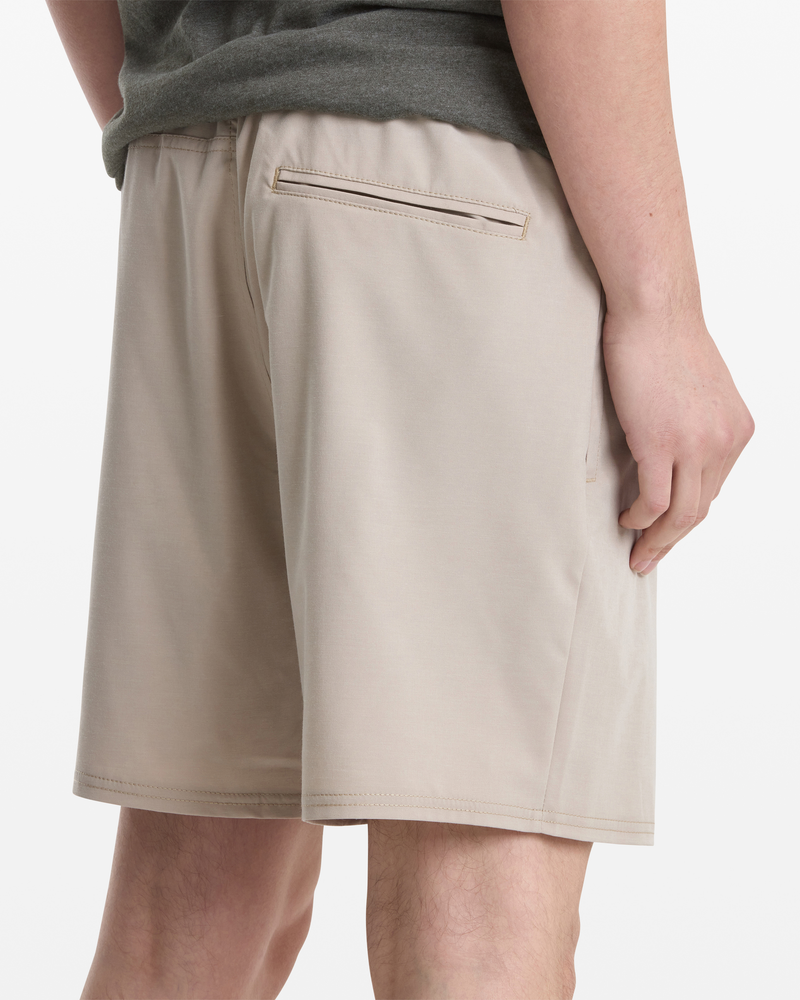 A person is shown wearing Volcom Men's Nomoly Elastic Waist Short 18 in light beige with a back pocket, standing with their hand by their side. The image also shows part of a gray shirt and the upper legs.