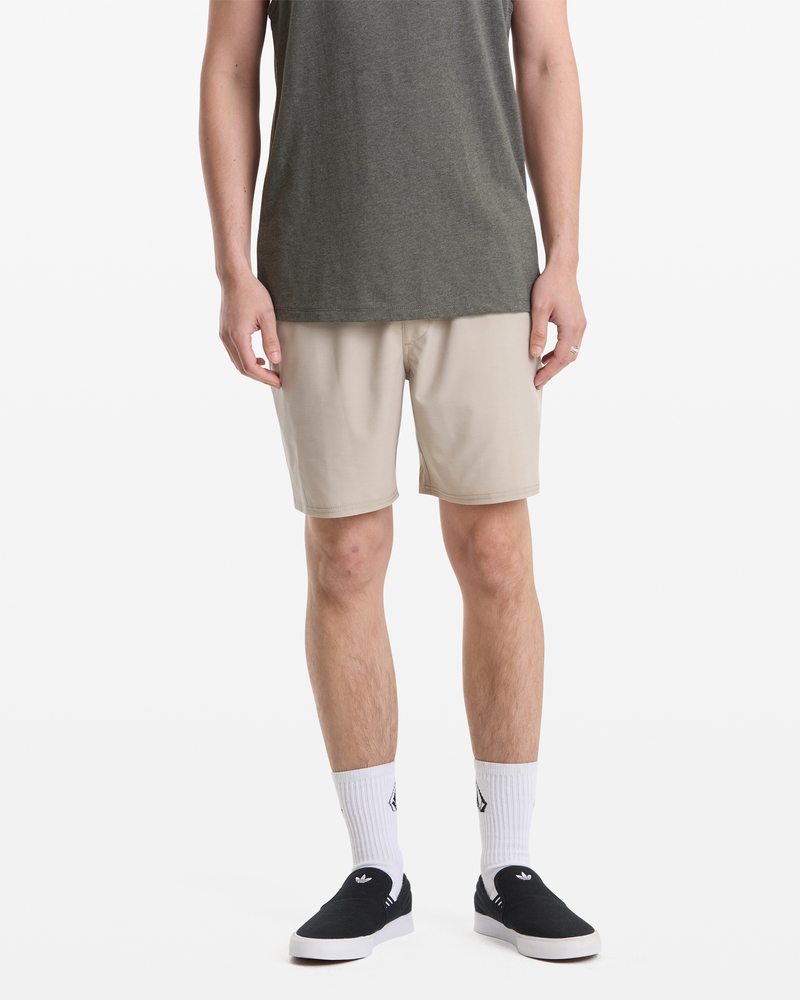 A man, shown from the shoulders down against a white background, wears gray tee, white socks, black slip-ons, and Volcom Men's Nomoly Elastic Waist Short 18 in light beige.