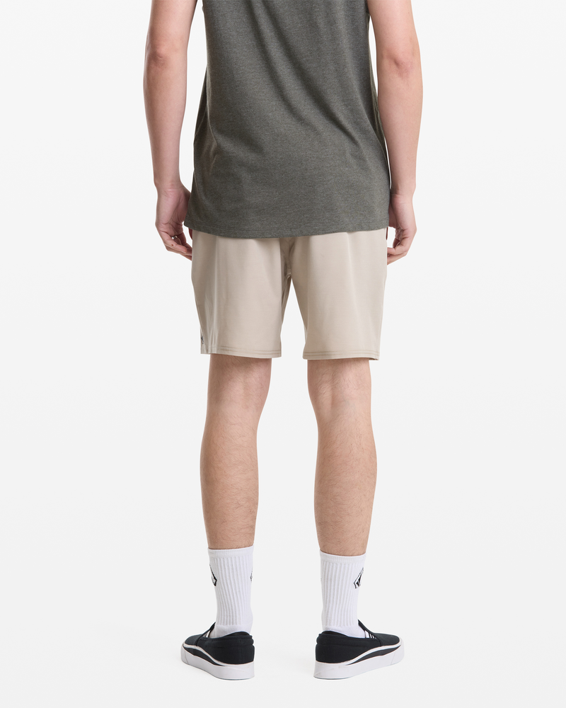 A person stands facing away, wearing a dark gray T-shirt, Volcom Men's Nomoly Elastic Waist Short 18 in beige, white socks with a small black design, and black-and-white sneakers against a plain white background.