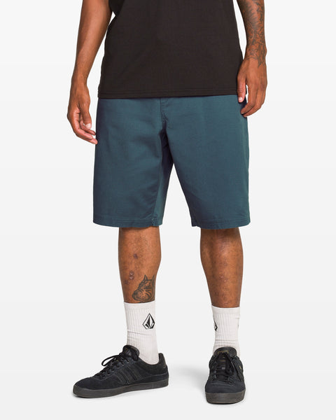 A person wearing Volcom Men's Freazy Loose Elastic Waist 22" Shorts in teal, a black t-shirt, white socks with a black logo, and black sneakers stands against a plain white background. Only the lower half of the body is visible.