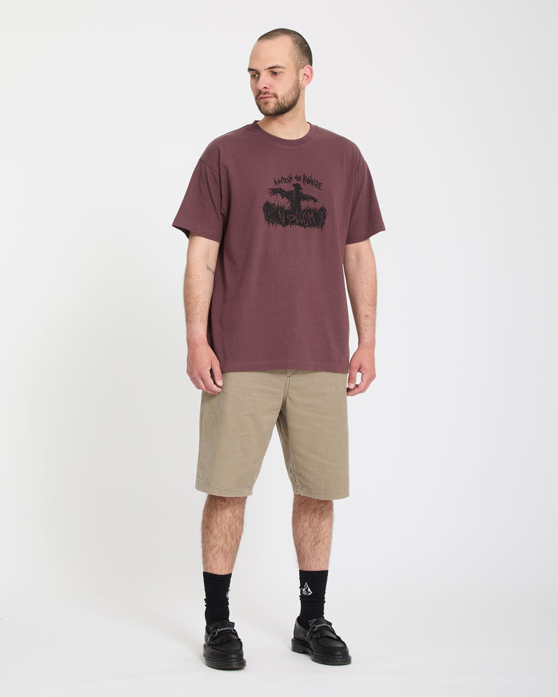 A bearded man stands against a white background, wearing Volcom Men's Freazy Loose Elastic Waist Short 22 in beige, a maroon graphic tee, black socks, and sneakers. He looks slightly left with a neutral expression.