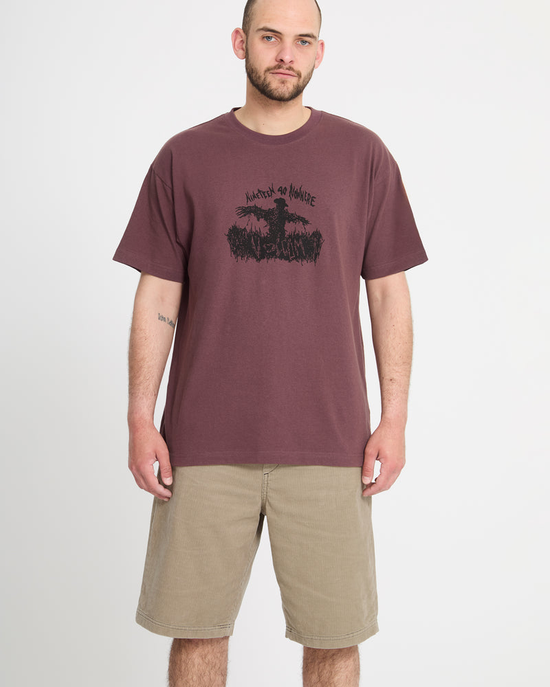 A man with short hair and a beard stands against a plain white background, wearing khaki Volcom Men's Freazy Loose Elastic Waist Short 22 and a maroon graphic tee. He faces forward, arms relaxed, with a neutral expression.