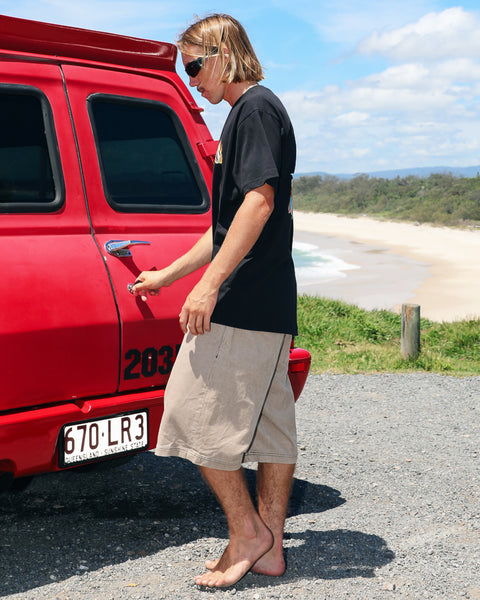 A man with long blond hair, sunglasses, a black t-shirt, and Volcom Men's Chillow 24" Shorts unlocks a red vehicle by the beach on a sunny day, with the ocean and sky in the background.