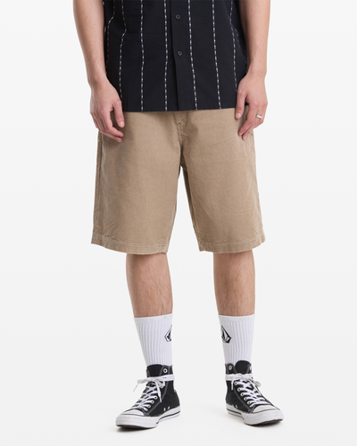 A person wears Volcom Men's Freazy Loose Elastic Waist Short 22 in beige, a black short-sleeved shirt with white stripes, white logo crew socks, and black low-top sneakers. The photo is cropped at the shoulders.