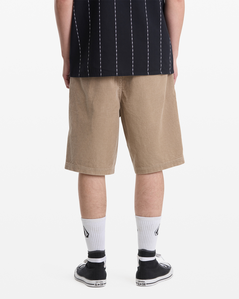 A person stands with their back to the camera, wearing a black and white striped shirt, Volcom Men's Freazy Loose Elastic Waist Short 22 in tan, white socks with a black logo, and black slip-on shoes.