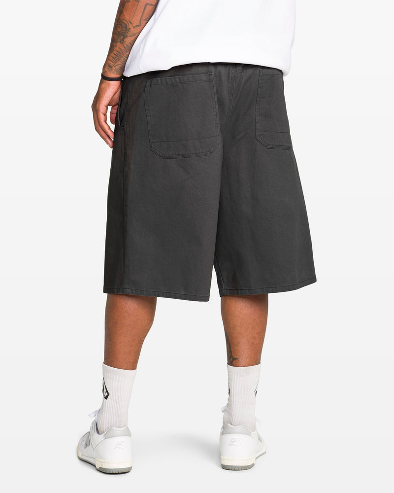 A person is shown from the back wearing a white t-shirt, white socks and sneakers, and Volcom Men's Chillow Plt Elastic Waist Chino 24" Shorts Vs in black. The person has a tattooed arm and stands against a plain white background.