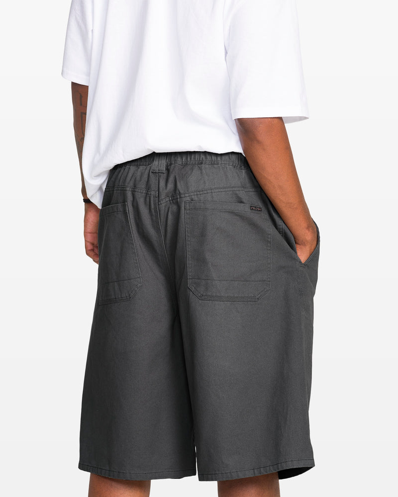 A man shown from the back wears a white t-shirt and Volcom Men's Chillow Plt Elastic Waist Chino 24" Shorts, standing with hands in both side pockets.