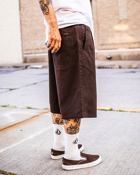 A person with leg tattoos stands on a sidewalk by a beige brick building, wearing a white shirt, Volcom Men's Chillow Plt Elastic Waist Chino 24" Shorts, white diamond logo socks, and brown slip-on shoes.