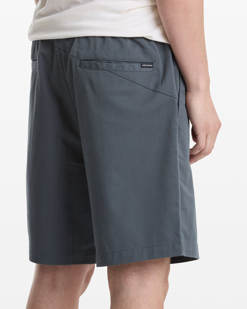 A person wearing a white shirt and Volcom Men's Frickin Elastic Waist Shorts in dark gray is shown from the back and side. The shorts feature a back pocket with a small rectangular Volcom label above it.