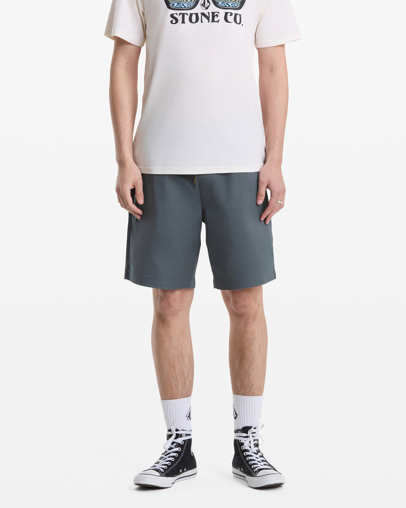 A person, head not visible, stands against a white background wearing blue-gray Volcom Men's Frickin Elastic Waist Shorts, a white graphic T-shirt, white crew socks, and black high-top sneakers.