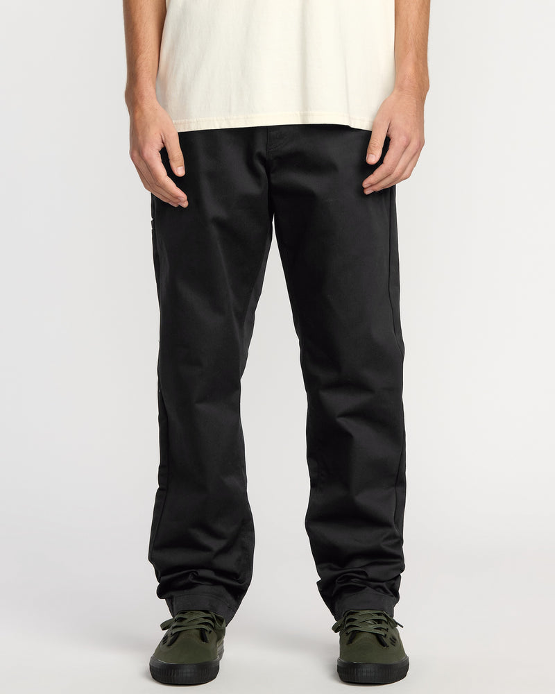 Only the lower torso, legs, and hands are visible as a person stands against a plain background wearing a white t-shirt, Volcom Men's Frickin Chino Pants in Dust Bowl Indigo, and dark green sneakers.