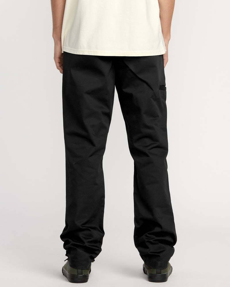 A man faces away from the camera, wearing a cream shirt, black Volcom Men's Frickin Chino Pants (relaxed fit), and black shoes against a plain white background.