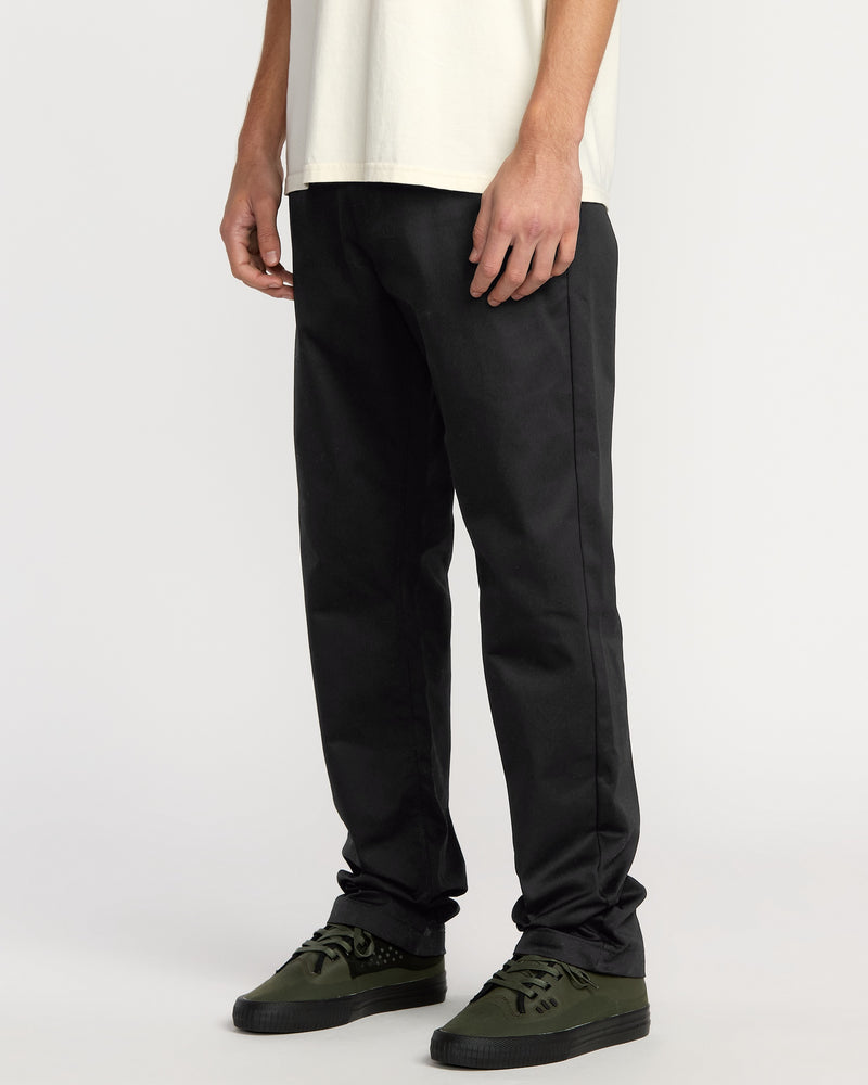 A person stands against a white background, shown from the torso down, wearing a white t-shirt, Volcom Men's Frickin Chino Pants in Dust Bowl Indigo (pre-laundered relaxed fit), and dark green sneakers.
