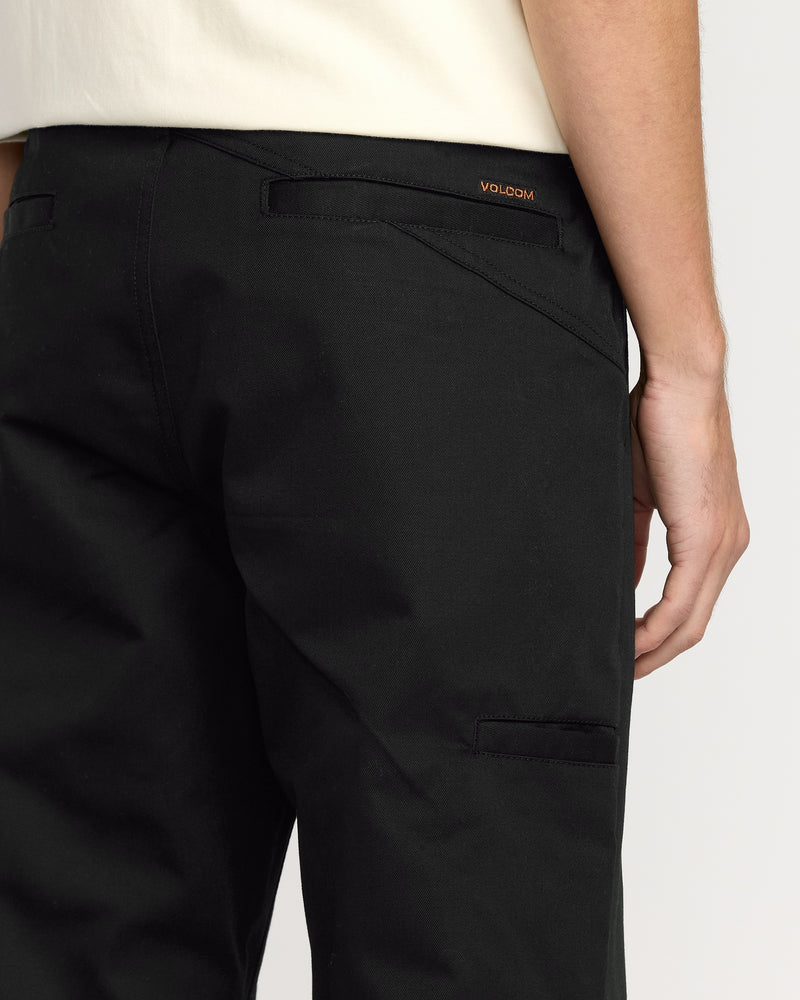 A close-up of a person wearing Volcom Men's Frickin Chino Pants in black with a relaxed fit, paired with a white shirt. The back and side pocket details are visible, featuring the small Volcom logo above the right back pocket.