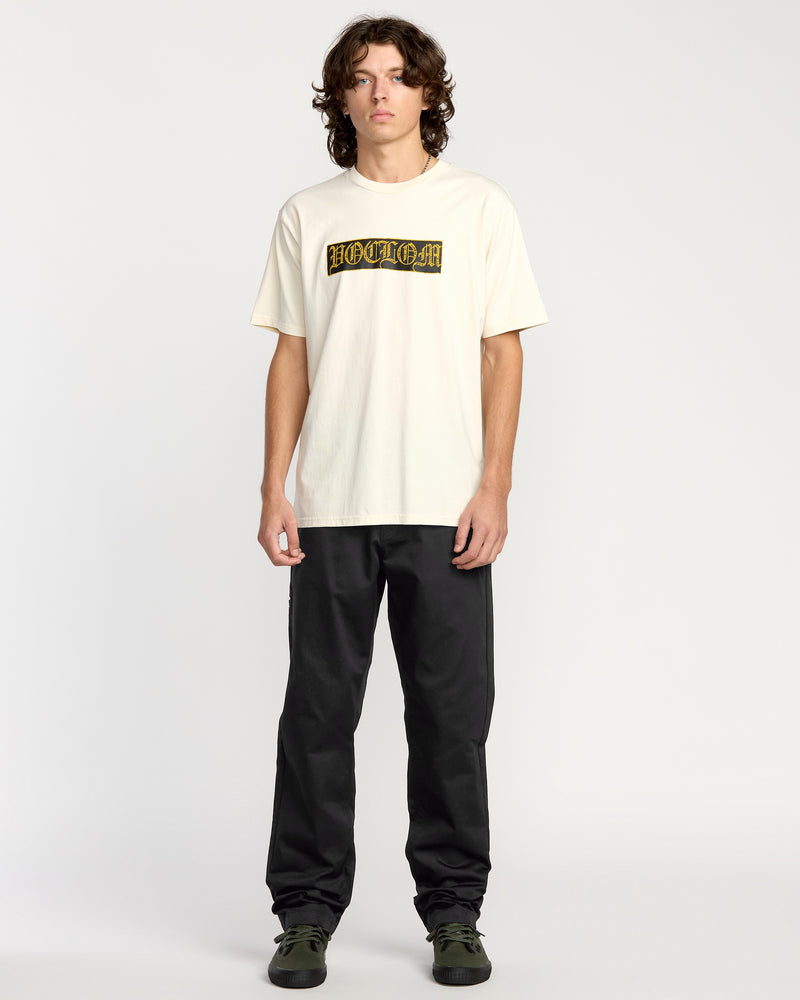 A young man with wavy brown hair stands against a plain background, wearing a cream pre-laundered HOODLUM T-shirt, Volcom Men's Frickin Chino Pants in black, and dark green sneakers.
