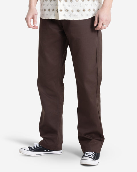 A person, face not visible, stands against a plain white background wearing Volcom Men's Frickin Chino Pants in dark brown, a patterned white shirt, and black low-top sneakers.