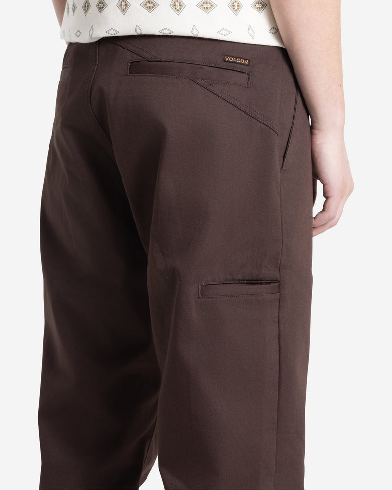 A person in Volcom Men's Frickin Chino Pants with relaxed fit, back and side pockets stands sideways, showing part of a cream diamond-pattern shirt. "Volcom" and "Frickin Chino Pant" branding appear above the back pocket.